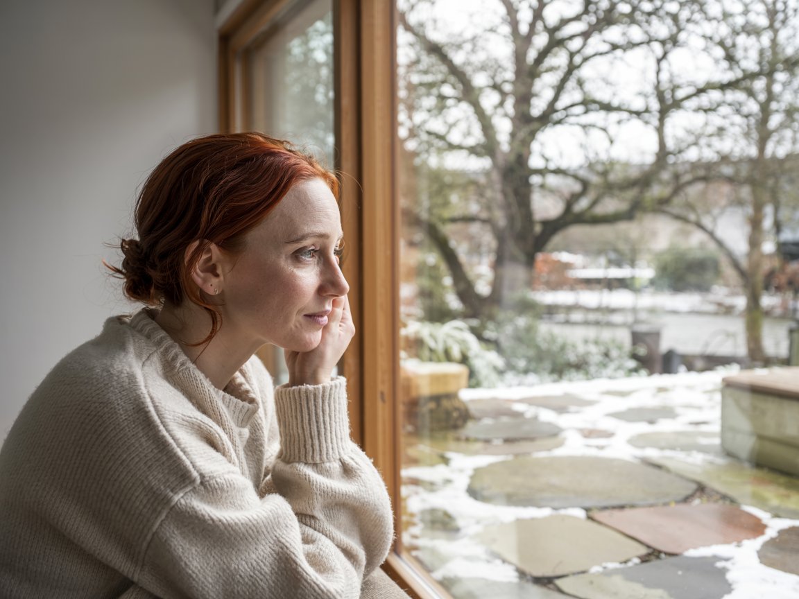 the interior glows with diffused white light — snow outside intensifies the reflection within. a woman in a heavy wool jumper leans against a timber-framed window, the glazing clear and near-invisible. rethink your windows 